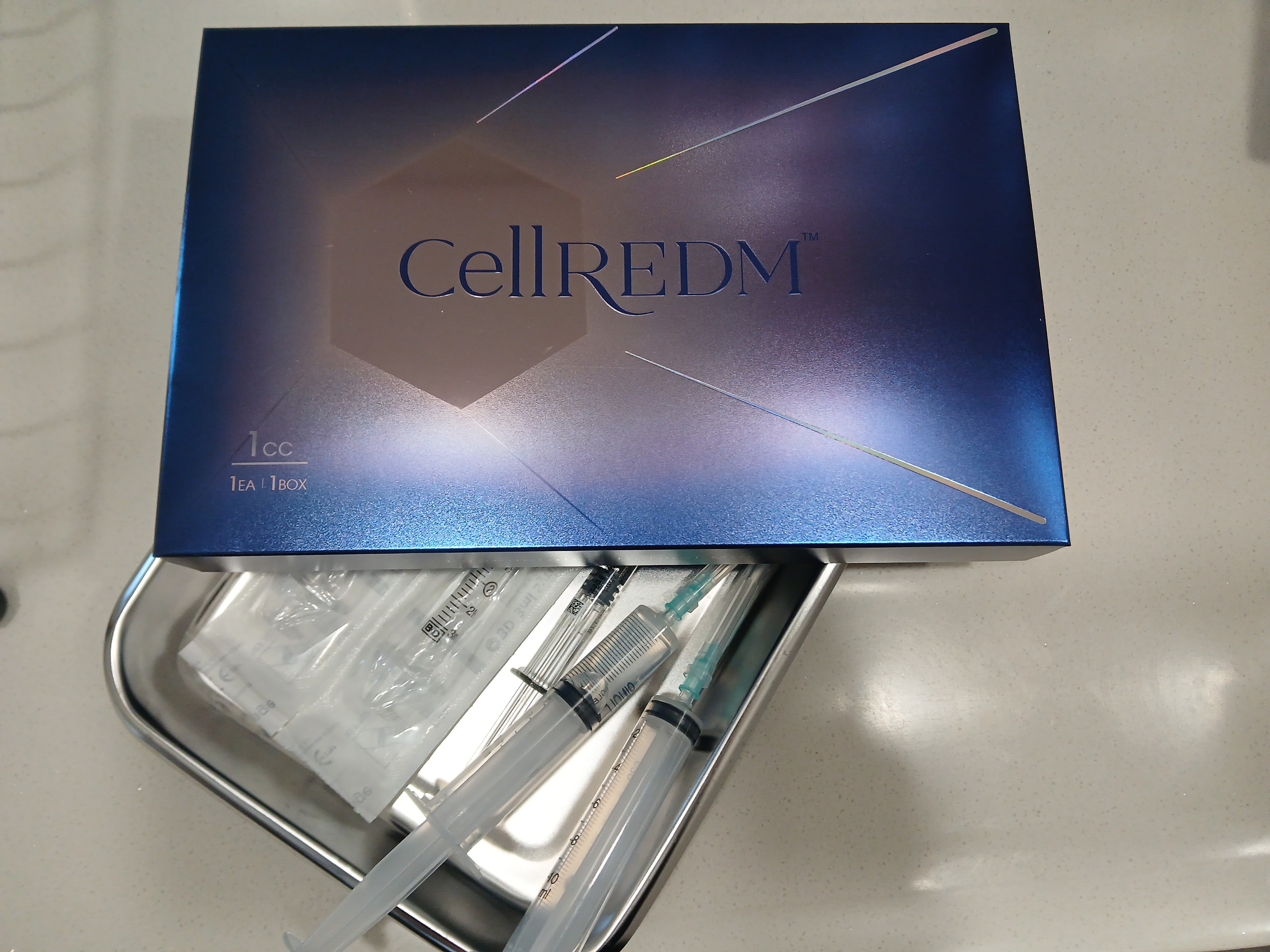 파란색 CellREDM 제품 상자 아래로 금속 트레이에 담긴 여러 개의 주사기와 의료용 도구들이 보이는 임상적인 분위기의 사진입니다.